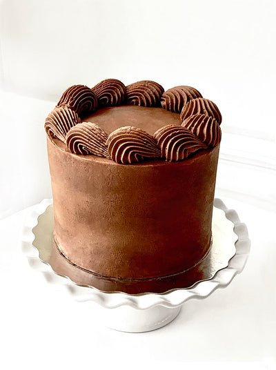 A chocolate cake, iced with mid-town brown icing is placed on a silver cake pad, which is sitting on a white cake stand with scalloped edges. The top of the cake has a large scale piped shell border around the circumference on top of the cake.