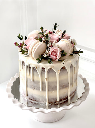 A Cake that has a thin layer of icing that has been scraped thin, sits on a silver cake tray that rests on a white scalloped cake stand. The Cake is covered in white chocolate ganache that drips down the sides leaving the naked cake exposed. The cake is topped with pink macarons and fresh roses and greenery. Gold foil flecks are artistically placed all over the cake 