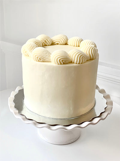 A cream-coloured iced cake sits on a silver cake pad which is resting on top of a white cake stand with scalloped edges. The top of the cake has a large scale shell border piped around the circumference in the same smooth creamy icing that covers the rest of the cake. 