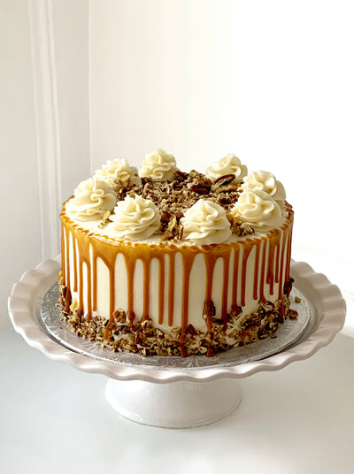 A wide, white iced cake is sitting on silver cake pad, on top of a white cake dish with scalloped edges. The cake has crushed pecans embedded on the lower quarter of the cake. Drips of caramel have been artistically placed around the edge of the cake, some of which are overlapping the nuts. The top of the cake has seven piped flower mounds of buttercream in the centre of these mounds are more crushed pecans - completely covering the top centre of the cake. 