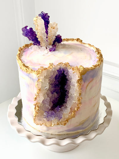 A white cake that has been painted with thin layers of purple, pink, and gold icing sits on top a silver tray that rests on a white scalloped cake stand. The cake has a chunk out of the side that is filled with purple and white candy crystals with and edge of gold around them and the top of the cake. It evokes an amethyst geode. The top of the cake has purple and white candy crystals that are stacked and spiked in on section to evoke more rock formations.