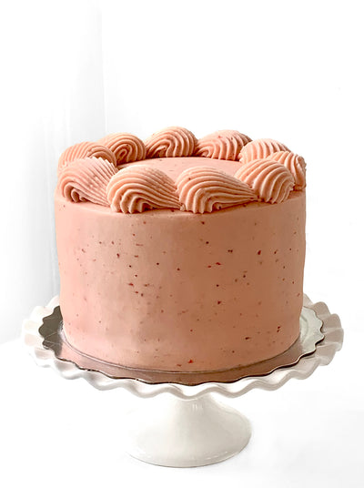 A light pink cake with red speckles sits on silver cake pad, which is on top of a white cake stand with scalloped edges. The circumference of the flat top of the cake has a large scale shell border, also piped in the pink icing. 